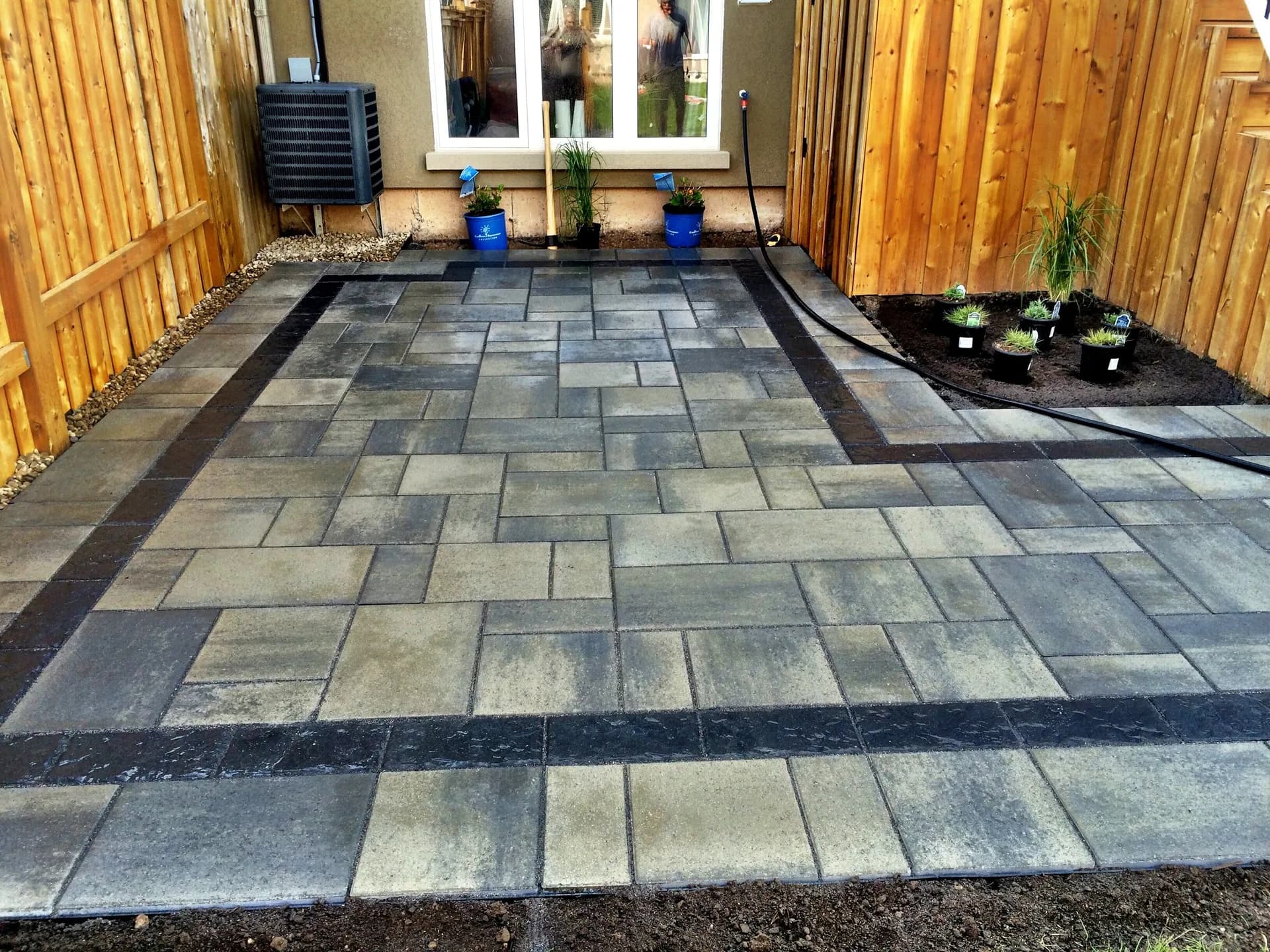 Backyard paver patio installation with decorative charcoal border inlay between wooden fences in Hamilton, Ontario