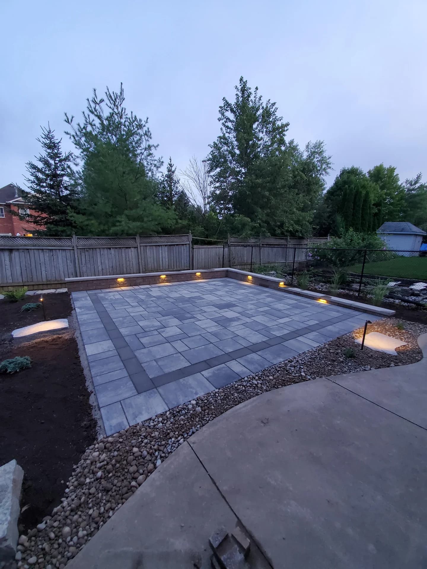 Backyard interlock patio with integrated step lighting, river rock border, and wooden fence at dusk in Dundas, Ontario