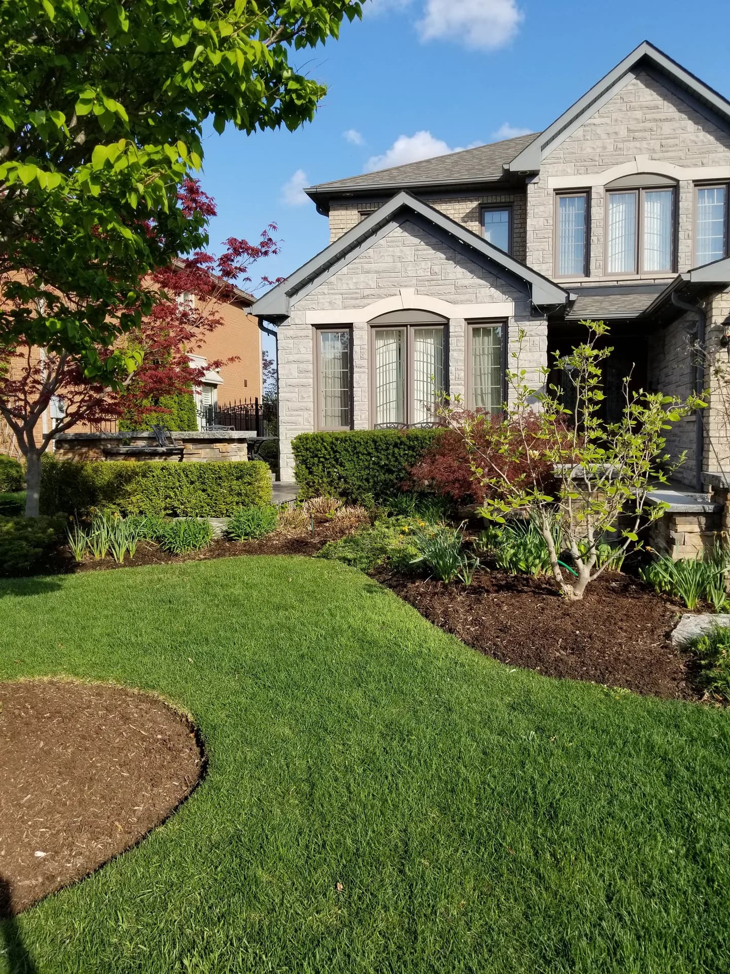 Front yard lawn mowing in Hamilton with fresh mulch beds, sharp lawn edging, and landscaped shrubs in front of a stone home